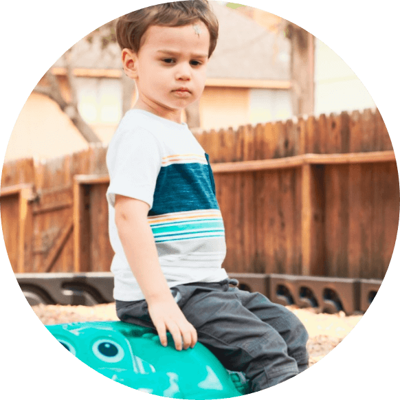 Boy with gray pants and striped t-shirt sitting on a green turtle seat on the playground while looking down to the ground
